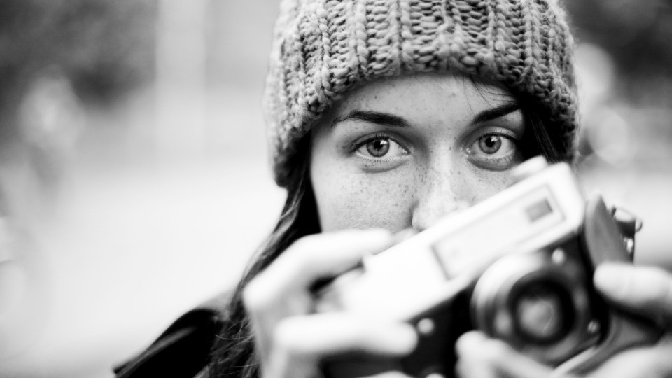 Black and white portrait of a photographer in a knitted hat raising a film camera to her eye, looking directly at the viewer over the top of it.